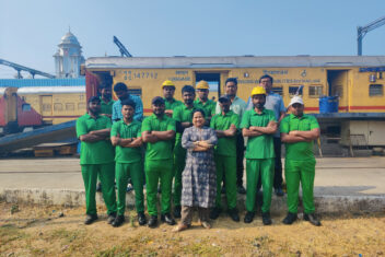 A group of men in green overalls with one woman in the middle in blue, all with their arms crossed standing in front of a train.