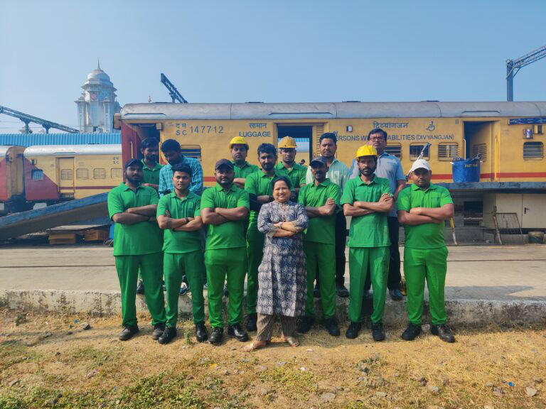 A group of men in green overalls with one woman in the middle in blue, all with their arms crossed standing in front of a train.